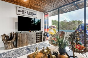 A living room with a television, wooden cabinet, decorative statues, a woven chair, and a view of the outdoors.