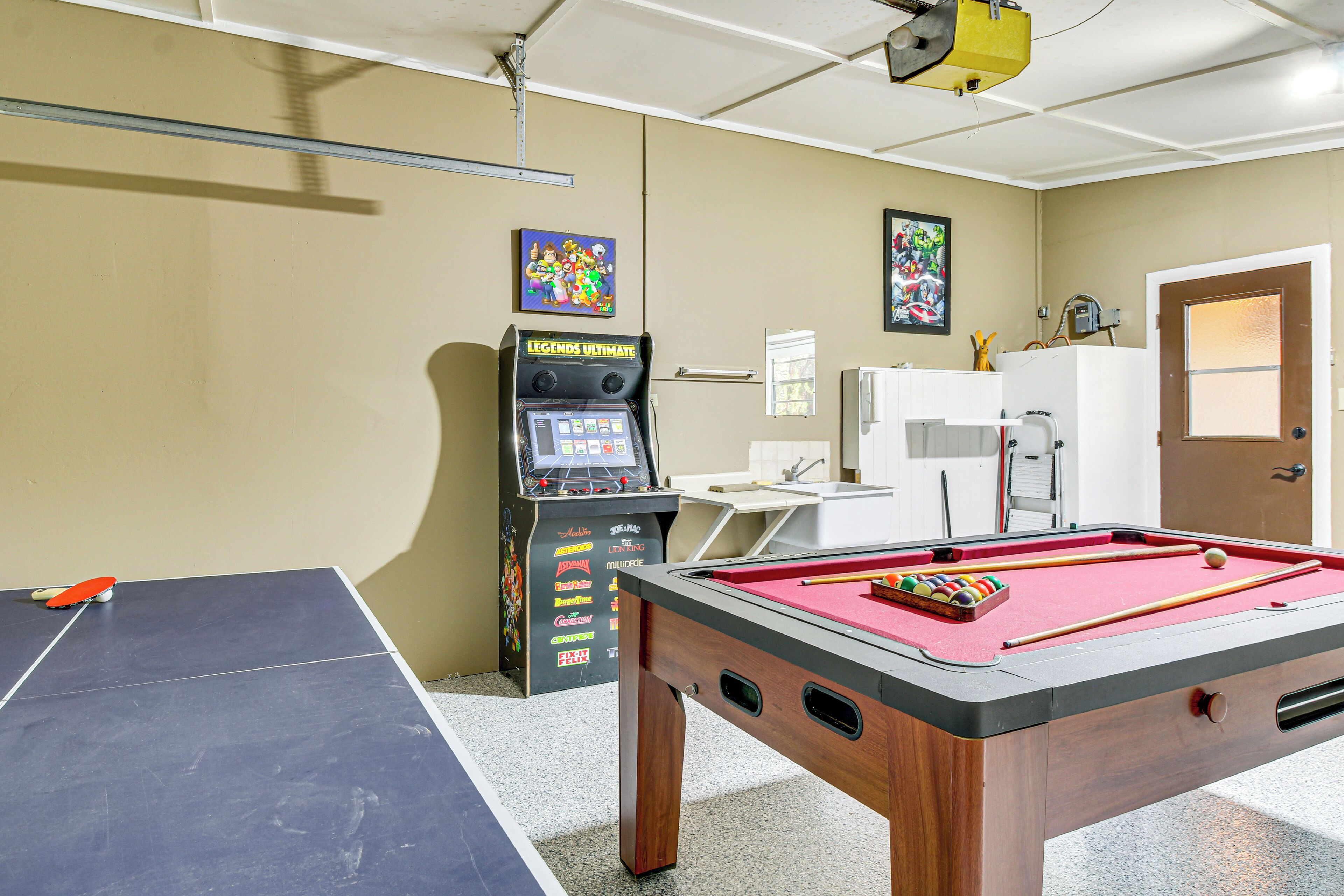 A pool table with a red felt surface, a foosball table, and a pinball machine in a room with beige walls and a door.