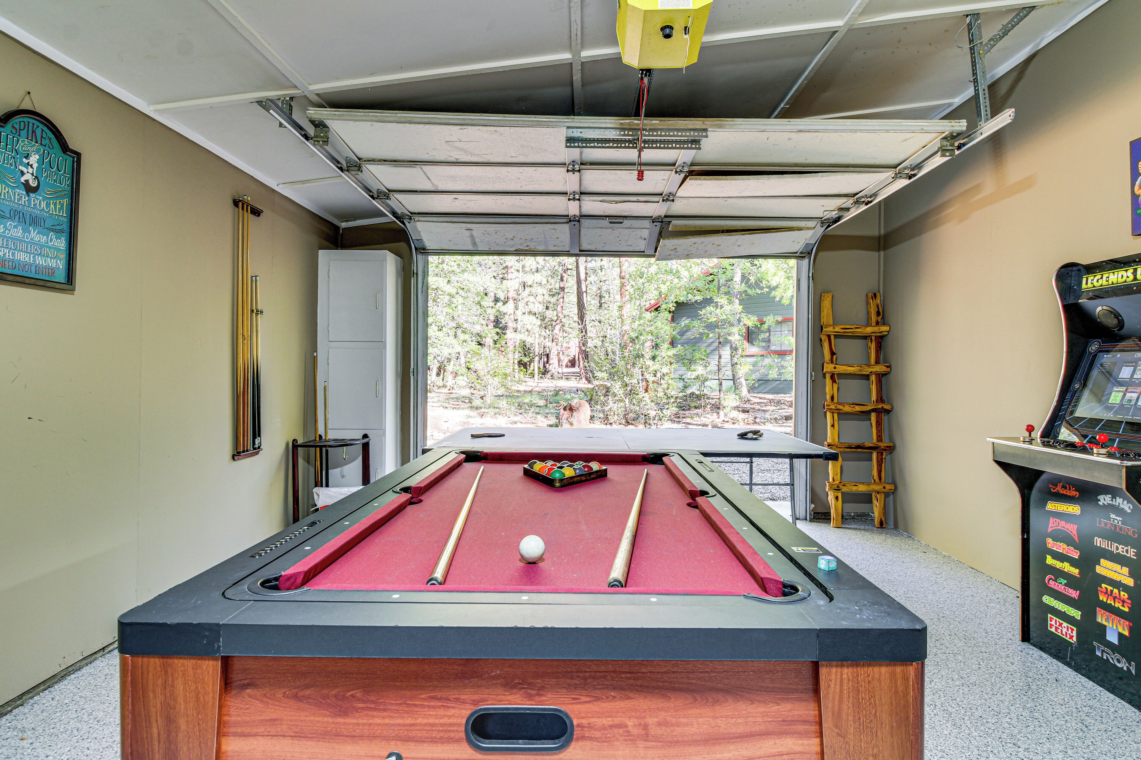 A pool table with a red felt surface, a cue ball, and a set of pool cues. There is a ladder leaning against the wall to the right of the table.
