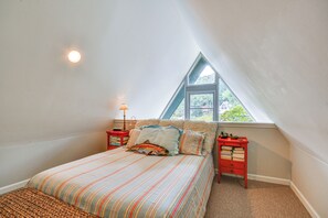 Um quarto com teto inclinado, uma cama com roupa de cama listrada, duas mesas de cabeceira com abajures e uma janela com vista para o verde.