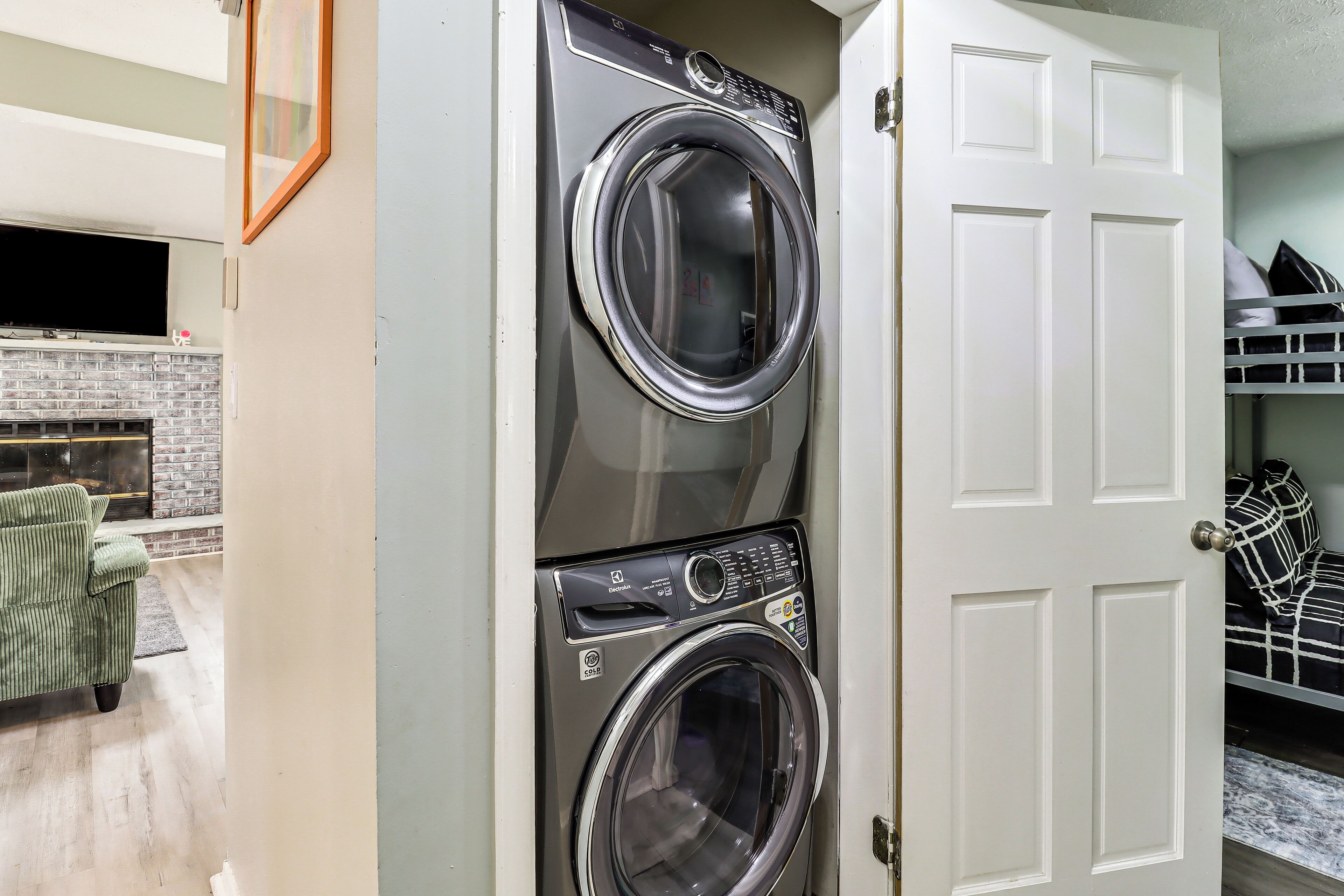 A laundry room with stacked washers and dryers, a door to a bedroom, and a brick fireplace.