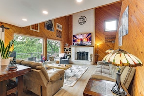 A cozy living room with a fireplace, wooden walls, a television, and a large window overlooking greenery. - Private Dock + Decks: Lake O' the Pines Escape (Jefferson)