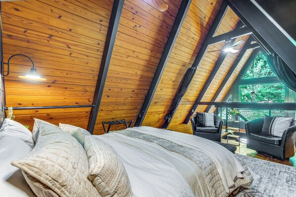 A cozy attic bedroom with a wooden ceiling, a bed with multiple pillows, a nightstand, and a view of the outdoors through a large window.