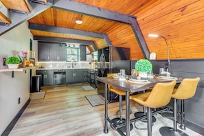 A modern kitchen with dark cabinets, a stainless steel refrigerator, and a dining area with a wooden table and yellow chairs. - Steps to Dale Hollow Lake! Cozy A-frame Cabin (Celina)