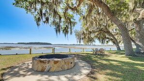 A stone fire pit surrounded by a concrete circle, a grassy area, and a view of a lake with trees and chairs.