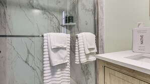 Un baño con paredes de mármol, puerta de ducha de vidrio, toallero con toallas blancas, un armario de madera y una botella de jabón corporal con aroma a coco y vainilla sobre el mostrador.