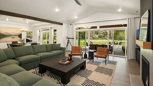 Een ruime woonkamer met een groene bank, een salontafel en een open haard. Er is een eethoek met een tafel en stoelen, en grote ramen met uitzicht op het landschap.