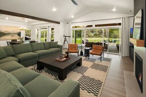 A spacious living room with a green sofa, a coffee table, and a fireplace. There is a dining area with a table and chairs, and large windows offering a view of the outdoors.