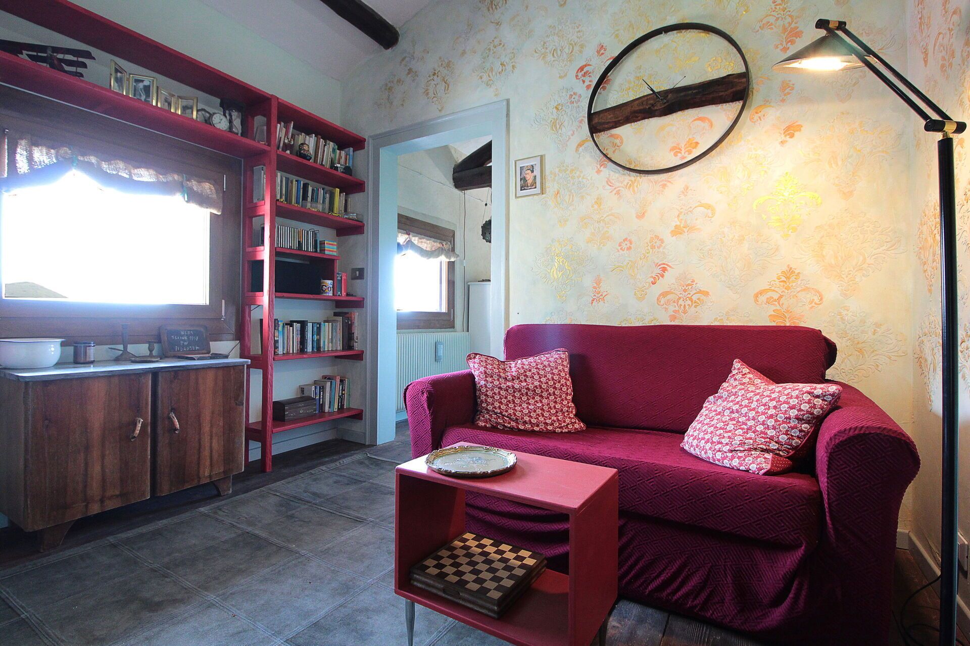 A cozy living room with a red sofa, a small side table with a chessboard, a bookshelf, and a wall-mounted lamp.