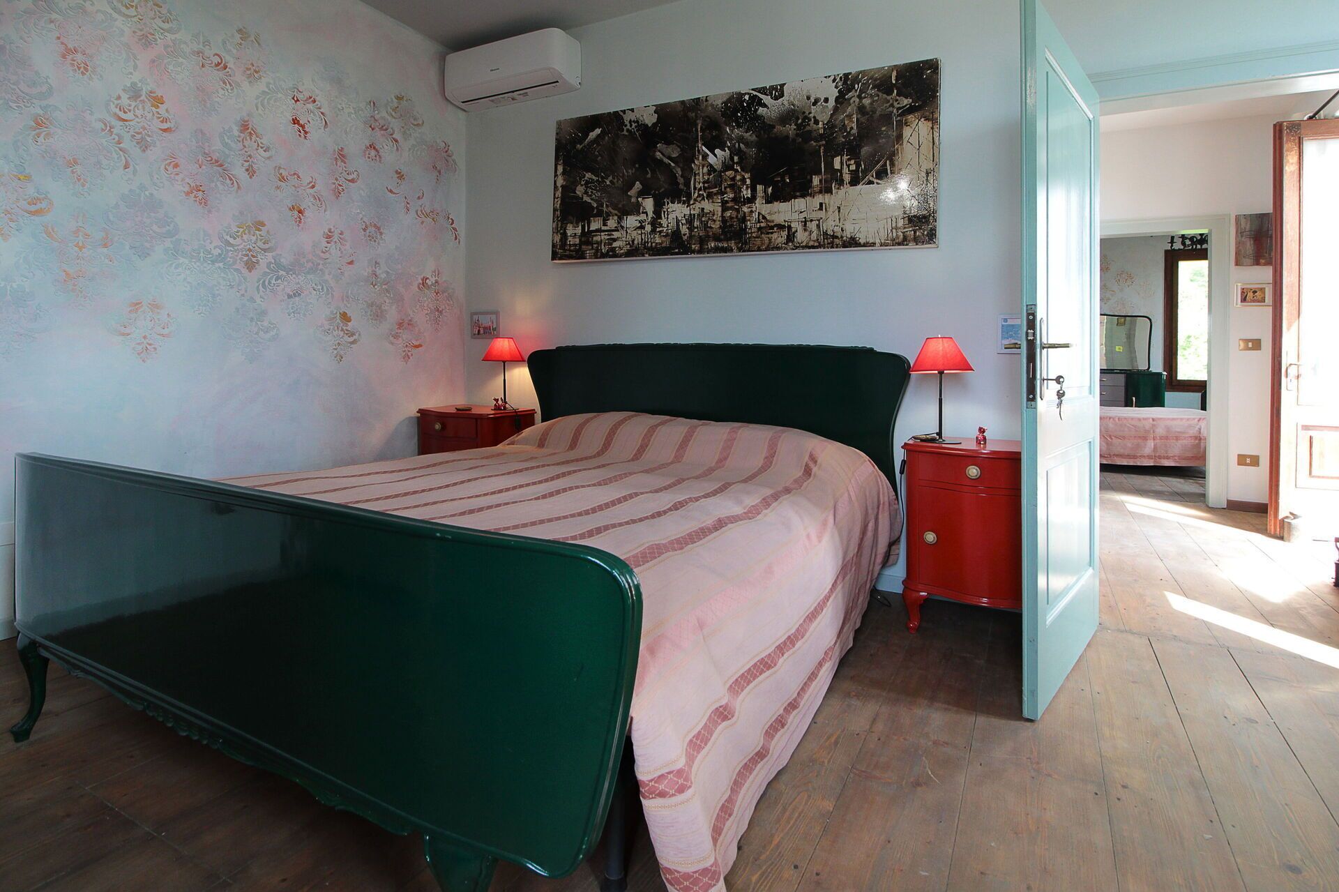 Una habitación con una cama de metal verde, dos mesitas de noche con lámparas, un aire acondicionado instalado en la pared y un cuadro abstracto grande.