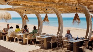 Een buitenterras met tafels en stoelen onder een rieten dak, met uitzicht op het strand.