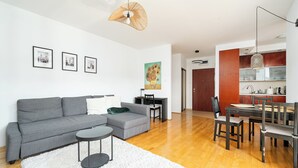 Una sala moderna con un sofá seccional gris, una pequeña mesa redonda y una alfombra peluda blanca. Se observa un comedor con una mesa y sillas de madera, y una cocina al fondo.