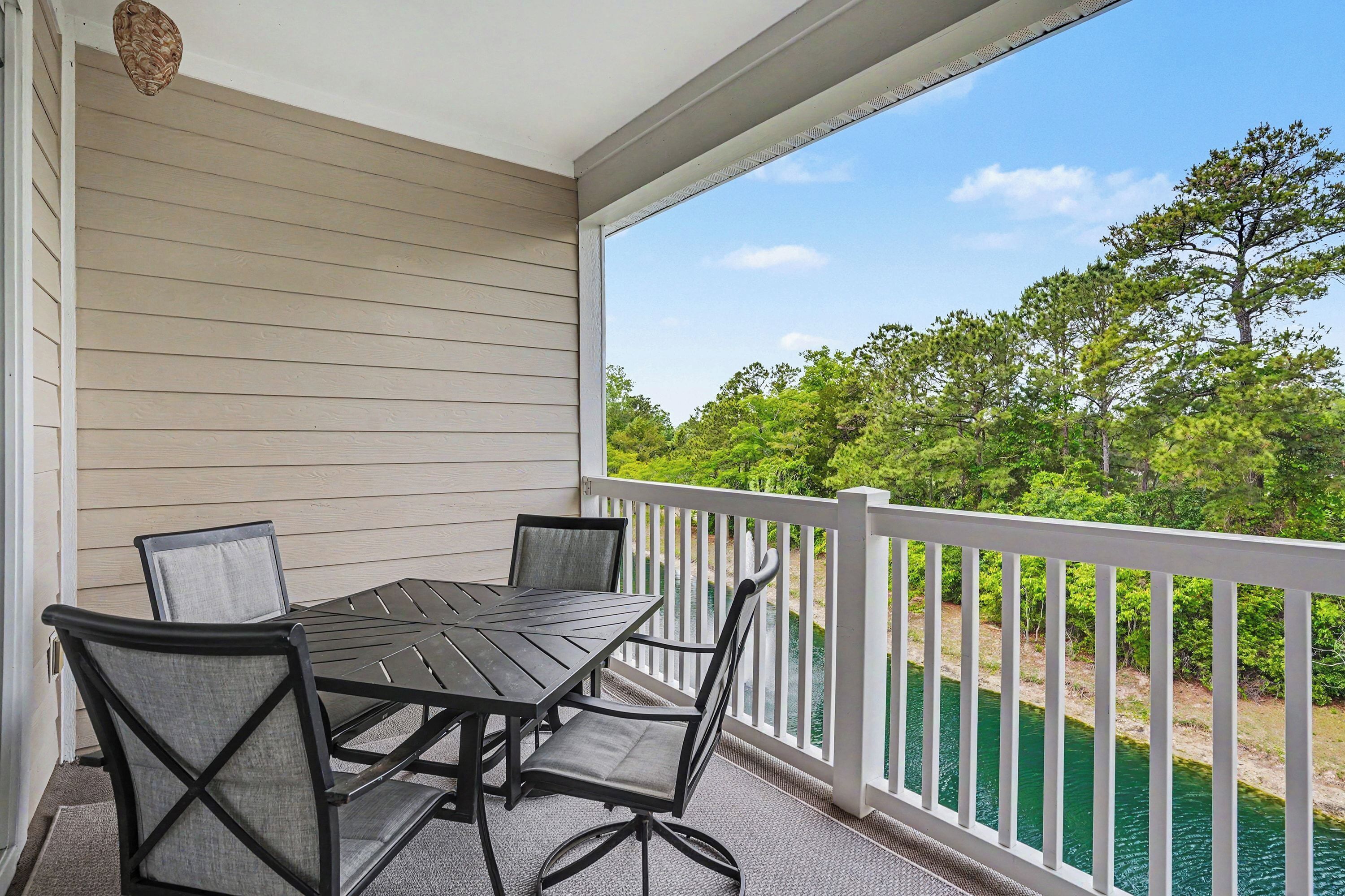 A covered outdoor area with a table and chairs, a white railing, and a view of a body of water and trees.