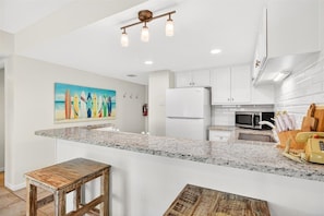 A modern kitchen with white cabinets, a granite countertop, a refrigerator, a microwave, and a painting on the wall. - Gulfstream Condos Unit 321 | Pool & Hot Tub | Private Balcony Gulf View (Corpus Christi)
