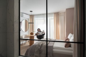 Ein Hotelzimmer mit Bett, Schreibtisch, Stuhl, rundem Spiegel und einem Fenster mit Vorhängen.