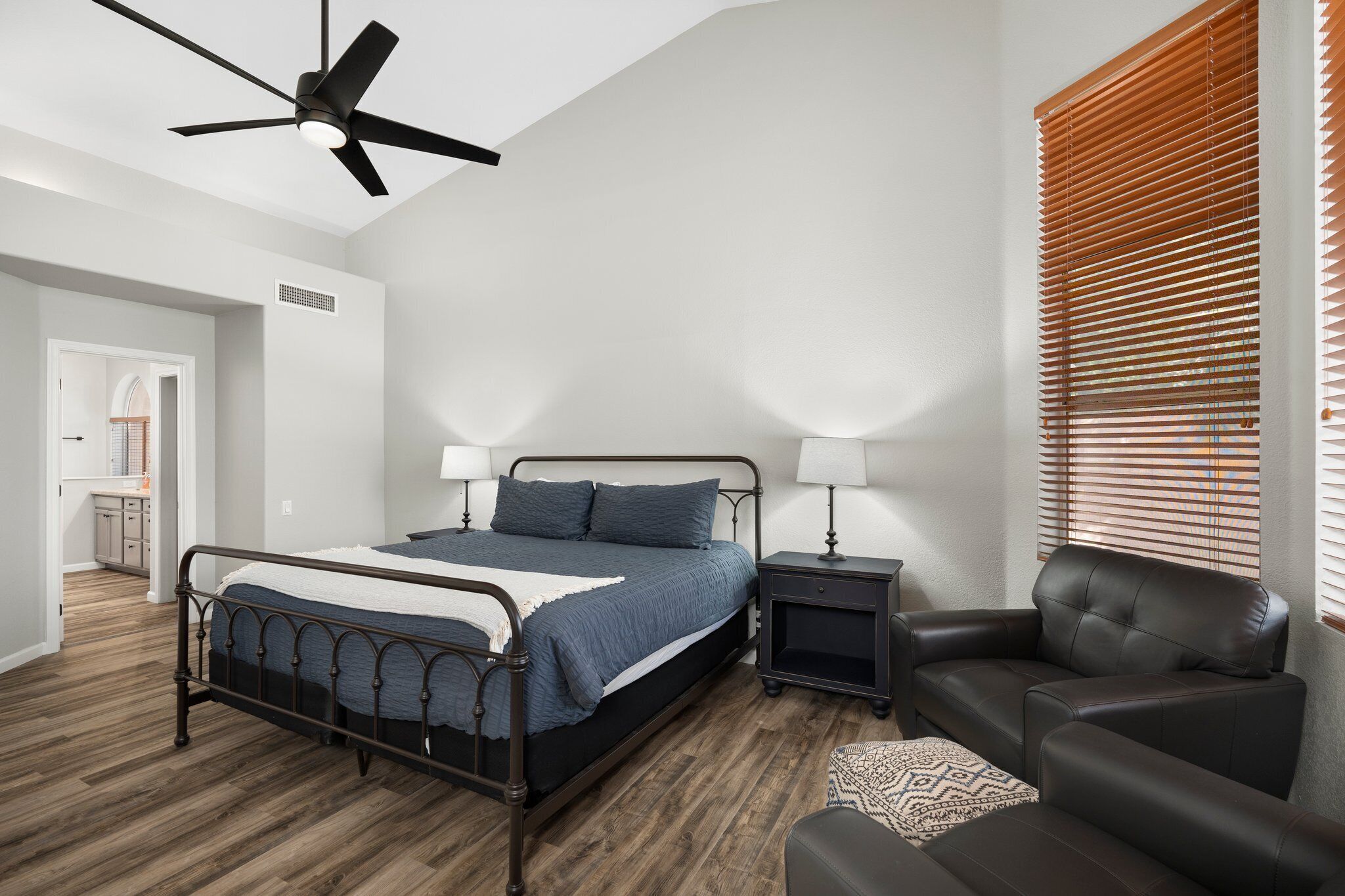 A bedroom with a bed, bedside tables, a chair, a ceiling fan, and a window with blinds.