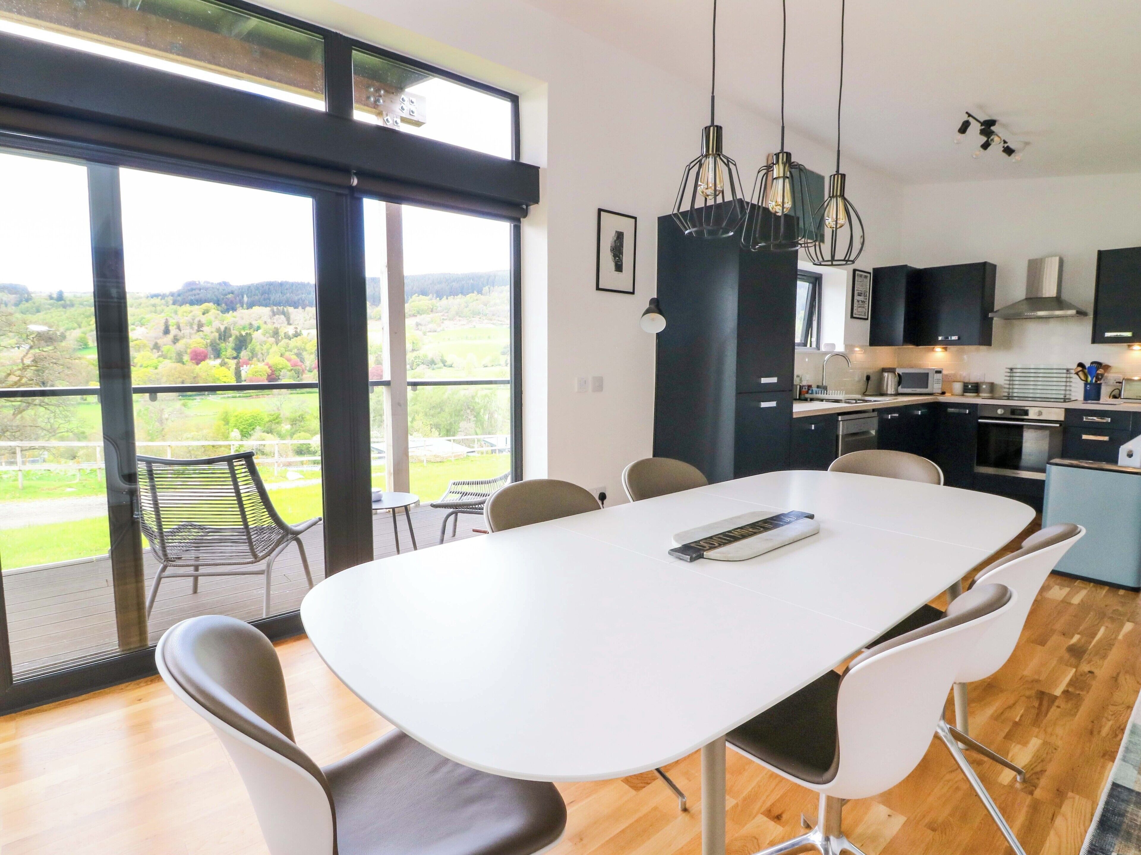 Una cocina moderna con una isla grande, un comedor con mesa y sillas blancas, y vistas a un paisaje verde a través de amplios ventanales.