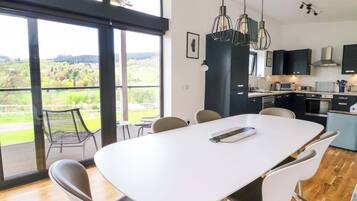 Una cocina moderna con una isla grande, un comedor con mesa y sillas blancas, y vistas a un paisaje verde a través de amplios ventanales.