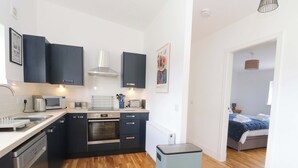 Cocina compacta con electrodomésticos modernos, un cesto de basura pequeño y vista hacia un dormitorio con cama y mesita de noche.