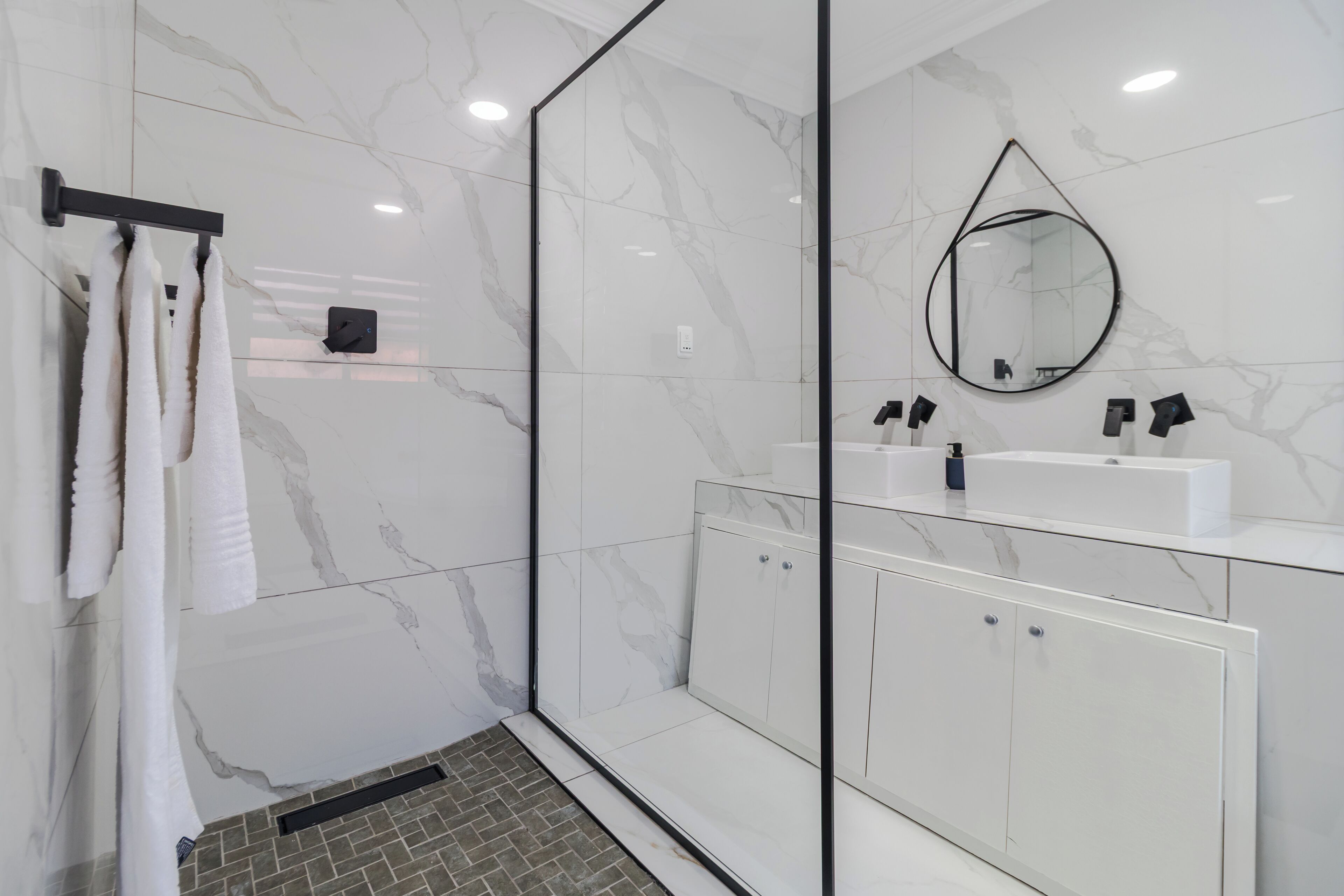 Ein modernes Badezimmer mit einer Duschkabine aus Glas, weißen Marmorwänden und einem weißen Waschtisch mit zwei Waschbecken.