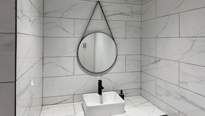 Un baño con azulejos de mármol blanco, un lavabo blanco y un espejo redondo con marco negro.