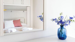 Een bed met witte beddengoed en een kussen met rode en witte strepen, een blauwe vaas met bloemen op een witte ondergrond, en een witte kast met zilveren handgrepen.