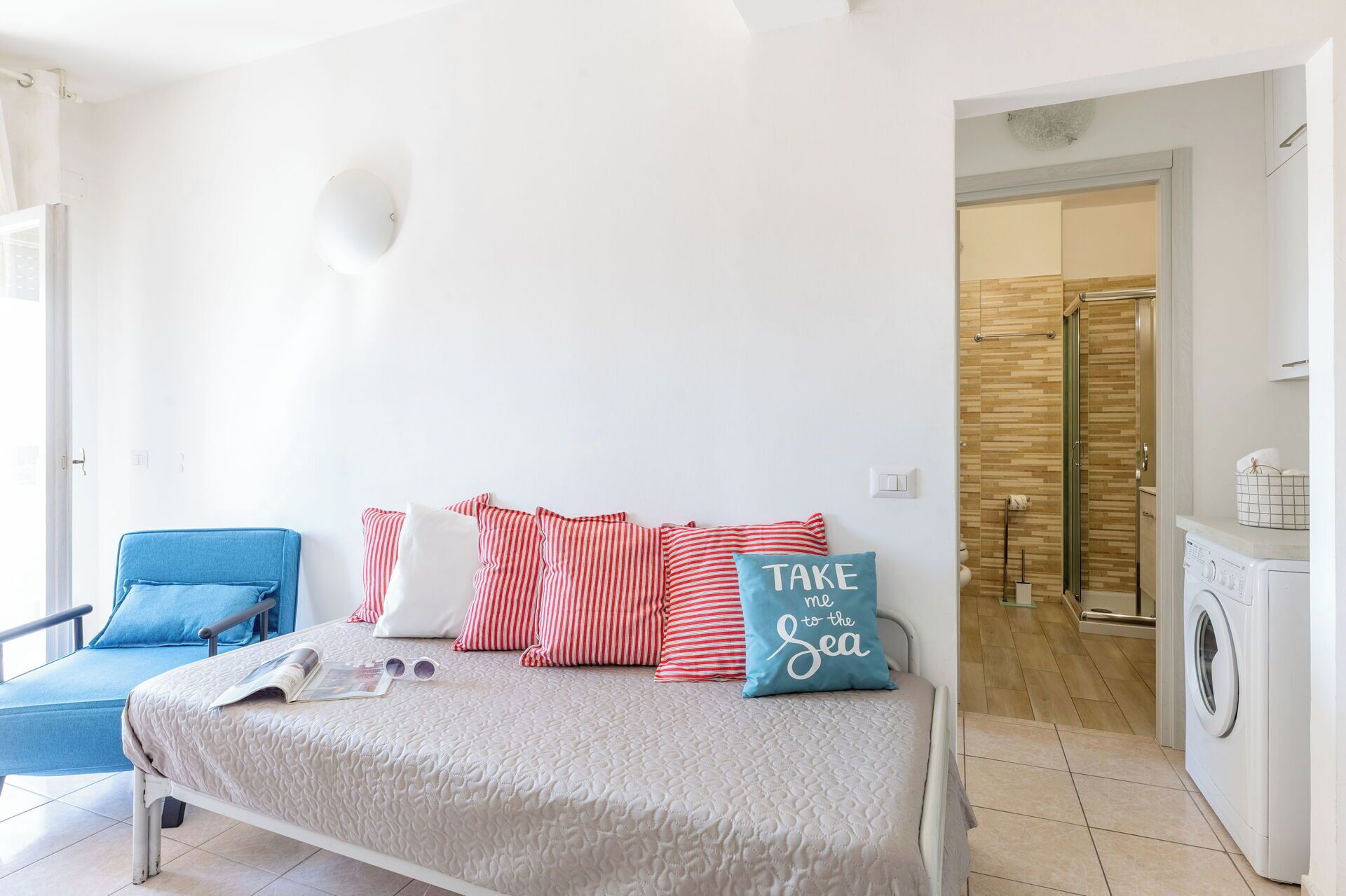 A bedroom with a bed, pillows, a chair, a washing machine, and a bathroom visible in the background.