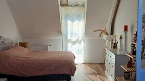 Une chambre à coucher avec un lit, un plancher en bois, une commode blanche, un luminaire suspendu et une fenêtre ornée de rideaux légers.