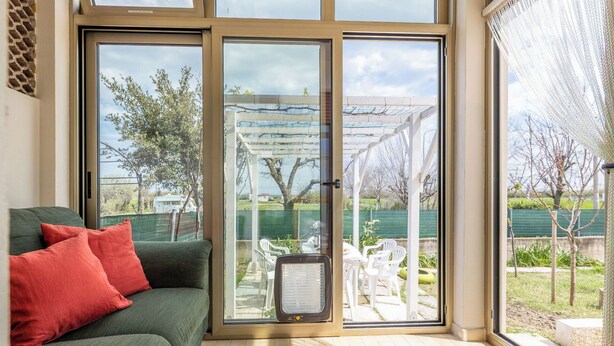 Una habitación con sofá verde, almohadas rojas, puerta de vidrio que da a un patio exterior con pérgola y una mesa con sillas.