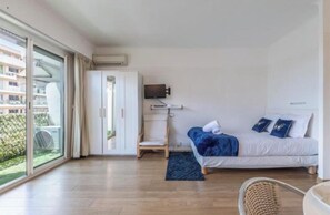 A modern hotel room with a large sliding glass door to the outside, a bed with blue bedding, a flat-screen TV mounted on the wall, and a wooden floor. - Apartment on the Croisette in Cannes (Cannes)