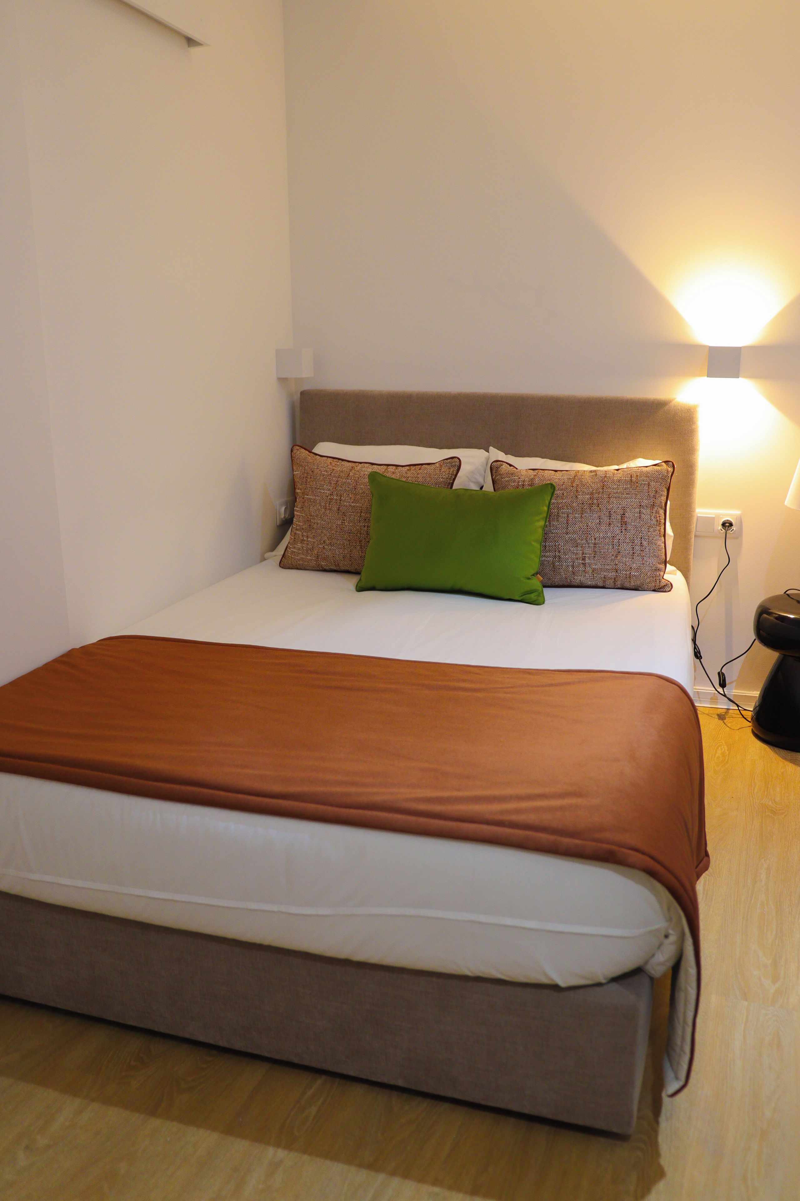 Una cama individual con una colcha marrón, un almohadón verde y dos almohadones estampados. Hay una lámpara de noche con pantalla blanca y un enchufe en la pared.