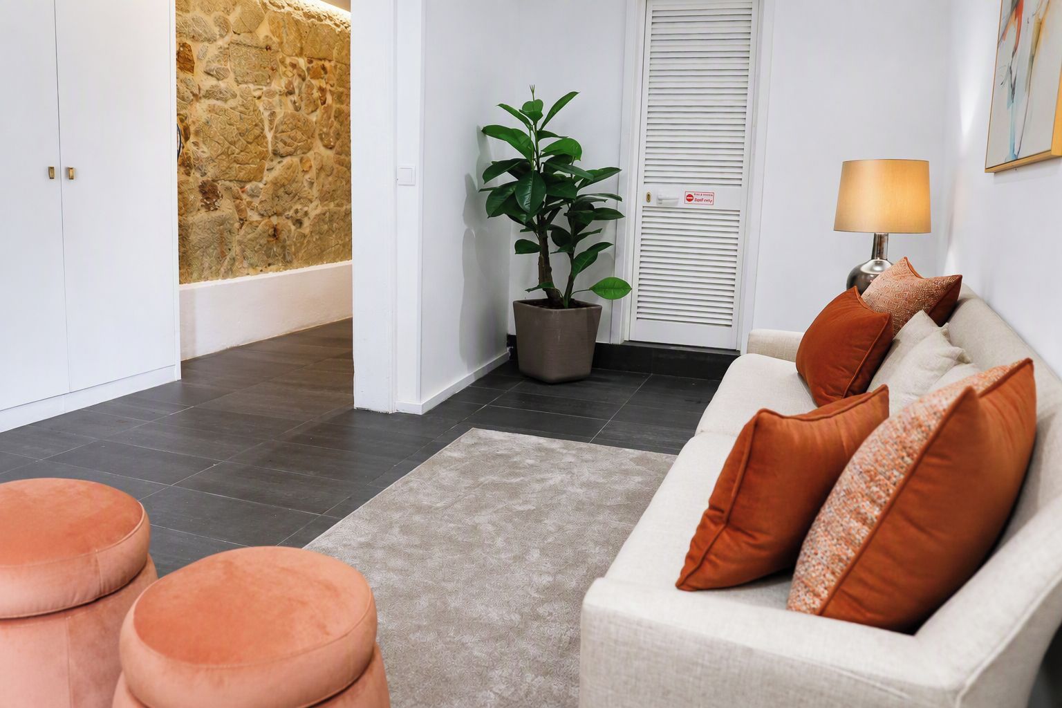 A modern living room with a sofa, ottomans, a potted plant, a lamp, and a stone wall.