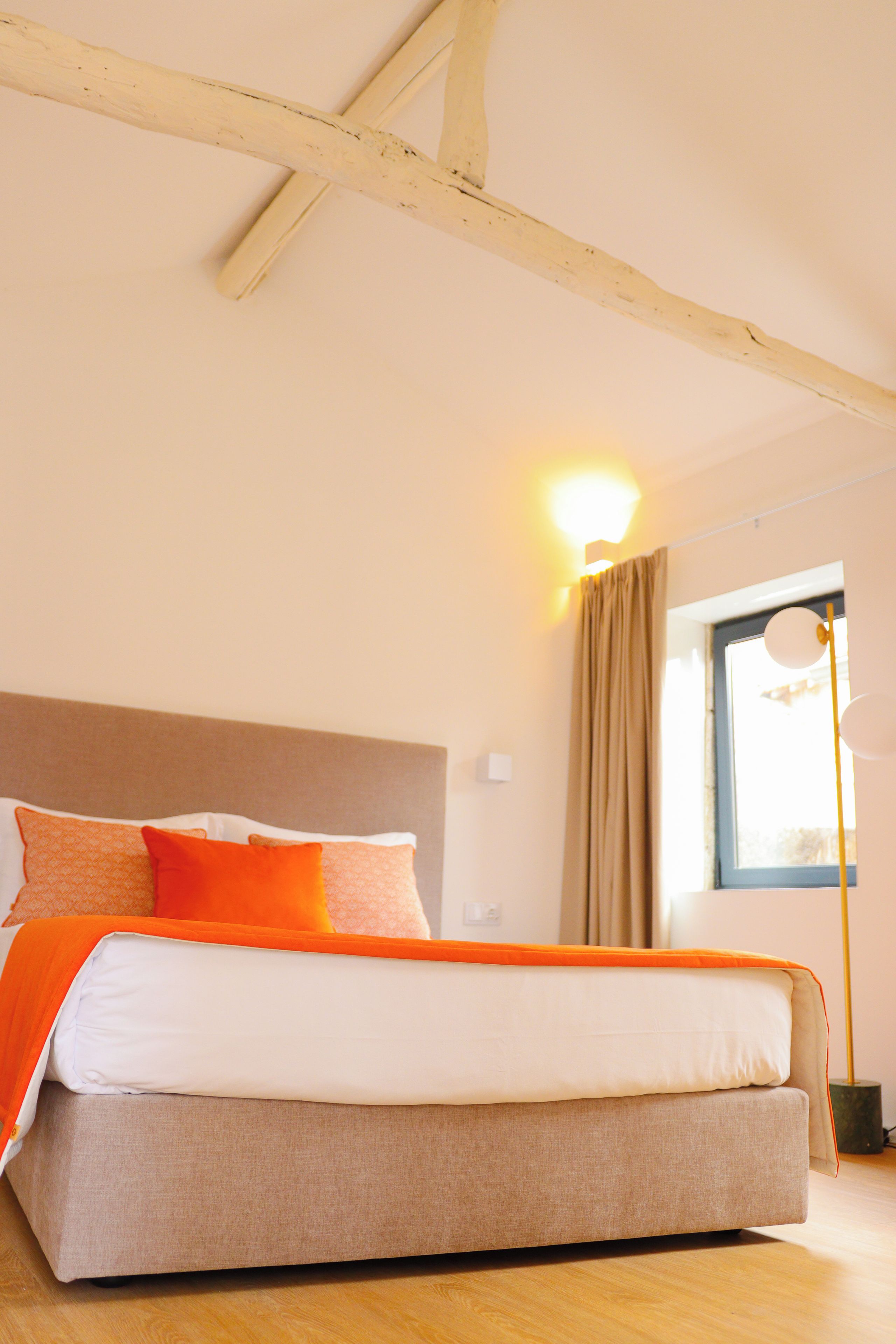 A bedroom with a bed, a nightstand, a lamp, a window with curtains, and a wooden ceiling.