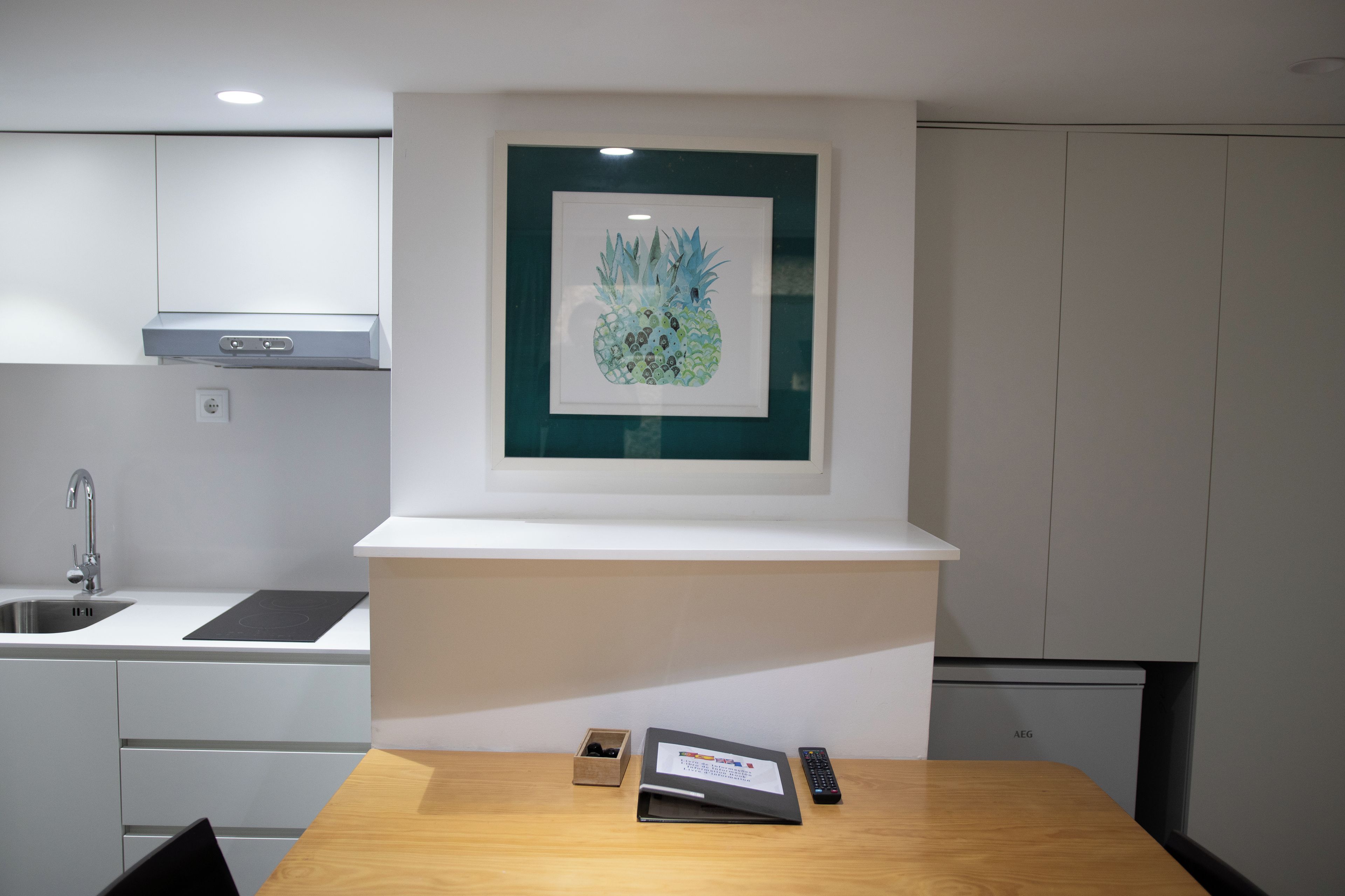 A modern kitchen with a white countertop, a framed pineapple artwork, and a wooden dining table with a remote control and a menu.