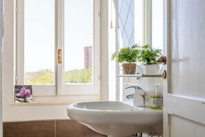 Ein Badezimmer mit einem weißen Waschbecken, einem Fenster mit Blick auf Grünflächen und einer Pflanze auf der Fensterbank.