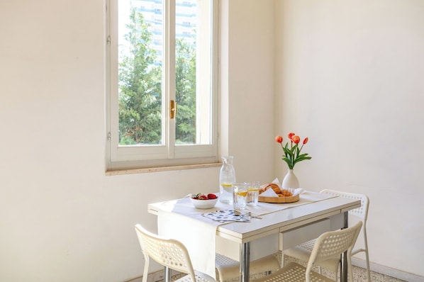 Ein Esstisch mit weißem Tischtuch, zwei weiße Stühle, eine Vase mit Blumen, eine Schale Erdbeeren, eine Karaffe und ein Teller Croissants.