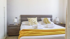 Een net opgemaakt bed met witte lakens, een gele plaid en meerdere kussens, waaronder een met een bladmotief. Twee nachtkastjes met lampen en een klok op één.