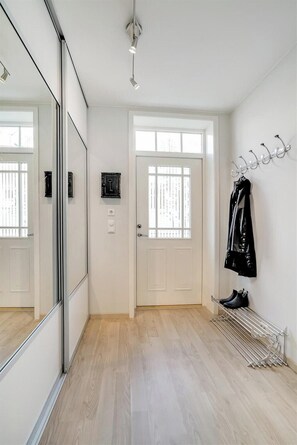 Ein Flur mit einer weißen Tür, einem Kleiderständer mit Jacken, einem Schuhregal und einem Spiegel, der Tür und Flur reflektiert.