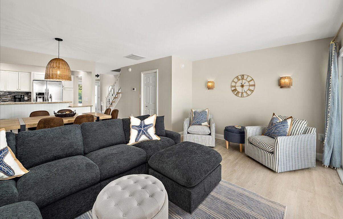 Een moderne woonkamer met een grijze hoekbank, een rond hockertje, een gestreepte fauteuil en een gestreepte bank. Er is een eethoek met een houten tafel en stoelen, een keuken op de achtergrond, en een trap die leidt naar een ander niveau.