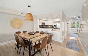 A modern kitchen with stainless steel appliances, a dining area with a wooden table and chairs, and a sign indicating a beach location.