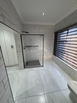 Ein modernes Badezimmer mit einer in Glas abgeschlossenen Dusche, weißen Marmorfliesen, einer Toilette und einem Fenster mit Jalousien.