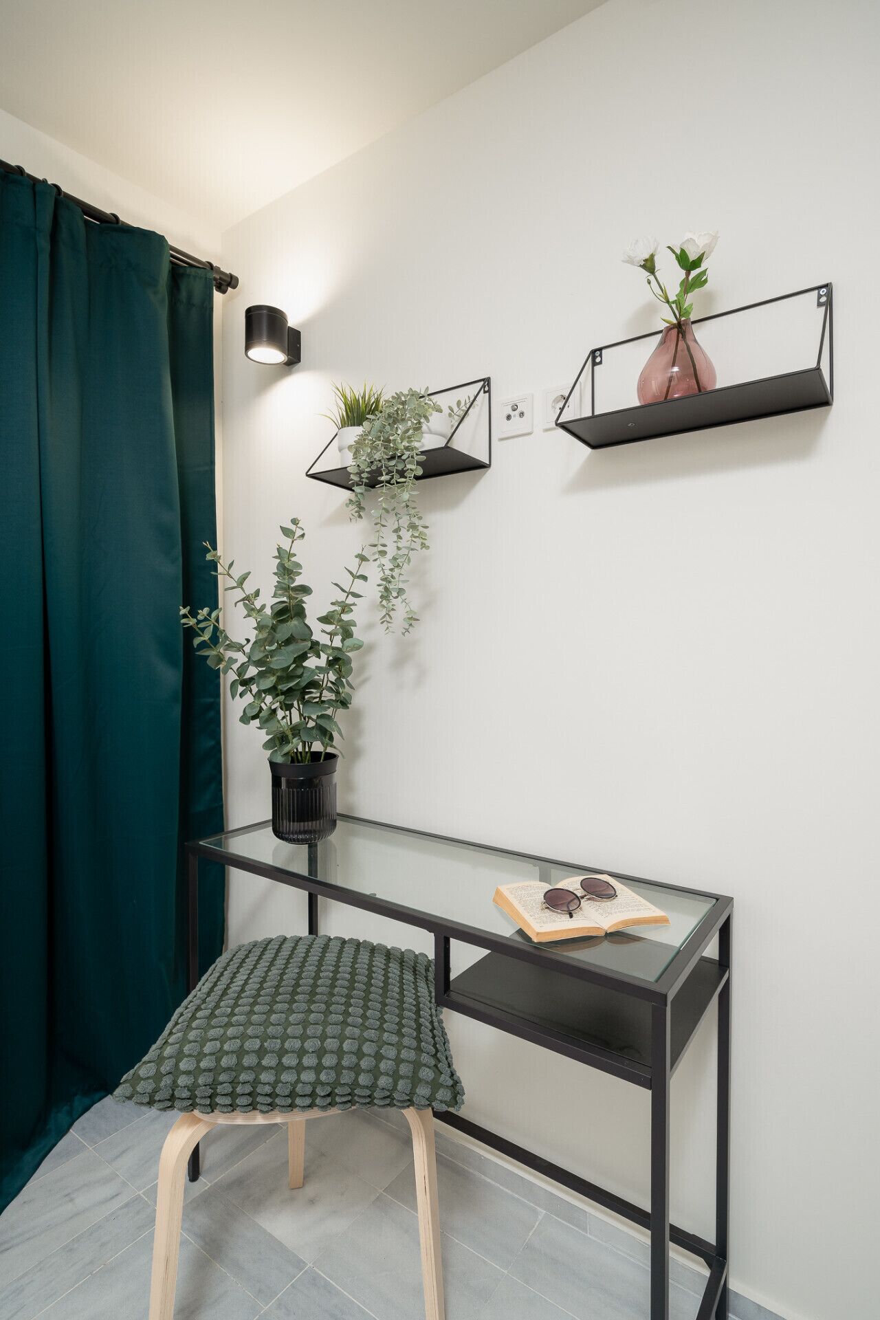 Ein Eckzimmer mit Schreibtisch, Hocker, Topfpflanze und einer an der Wand befestigten Ablage mit Pflanze.