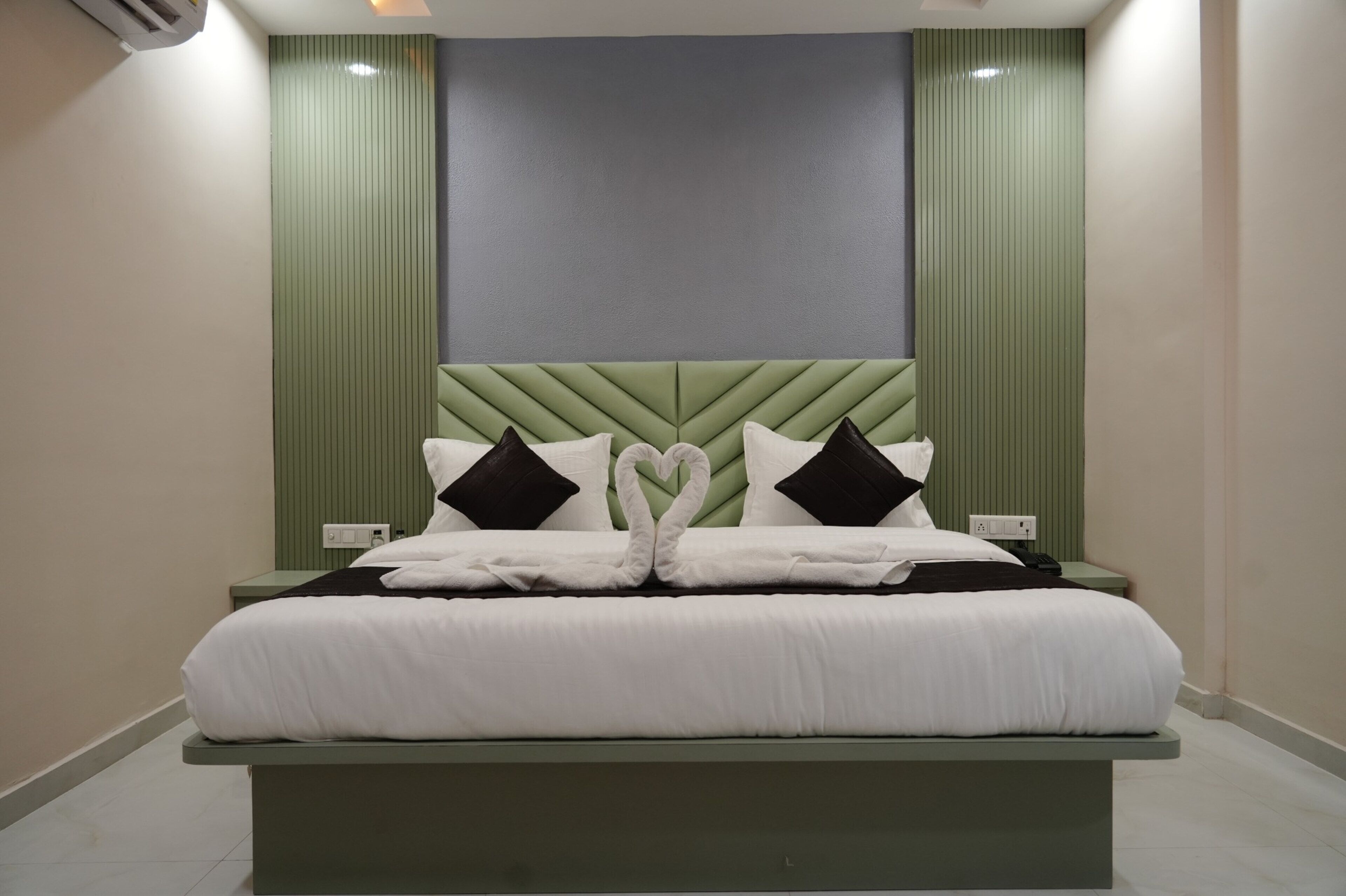 Una cama con una toalla dispuesta en forma de corazón, un cabecero verde y una pared en tonos neutros.