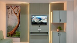 A modern living room with a flat-screen TV mounted on a wall, a large tree painting, a green sofa, and a cabinet with a plant.