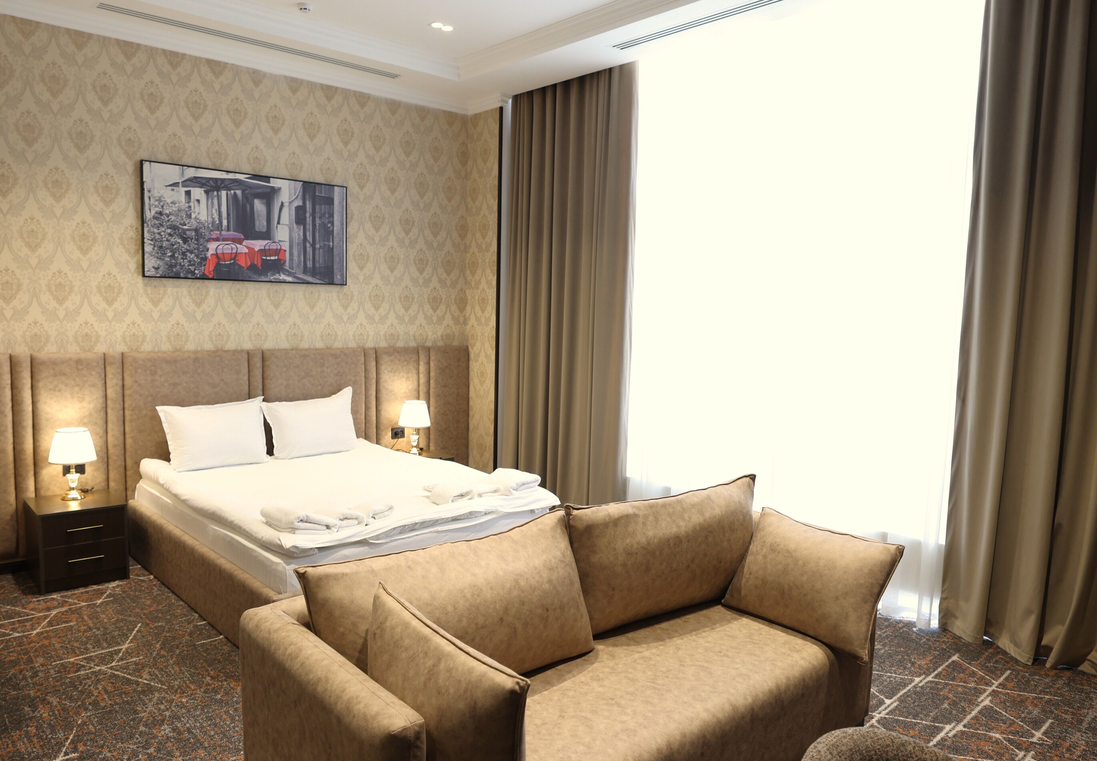 Hotelkamer met een bed, twee nachtkastjes met lampen, een bank, een behangen muur en een raam met gordijnen.