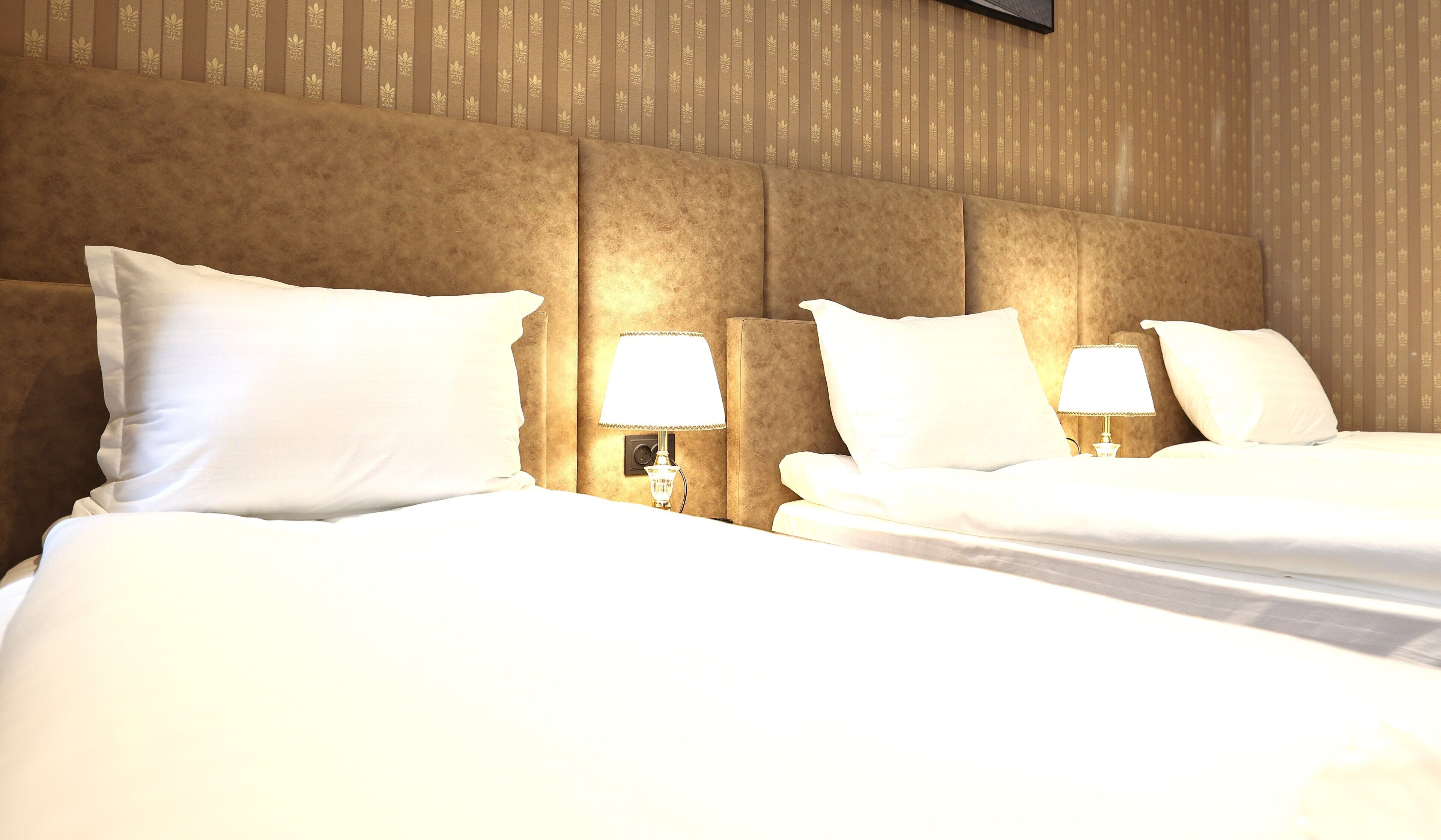 A hotel room with two beds, each with white bedding and pillows, and bedside lamps. The room has a textured wall with a vertical pattern.