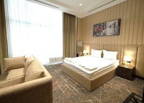 Una habitación de hotel con una cama grande, un sofá, una mesita de noche con lámpara, un televisor montado en la pared y una alfombra estampada.