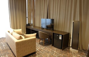 Habitación de hotel con sofá, escritorio con televisor de pantalla plana, silla, mini refrigerador y cortinas.