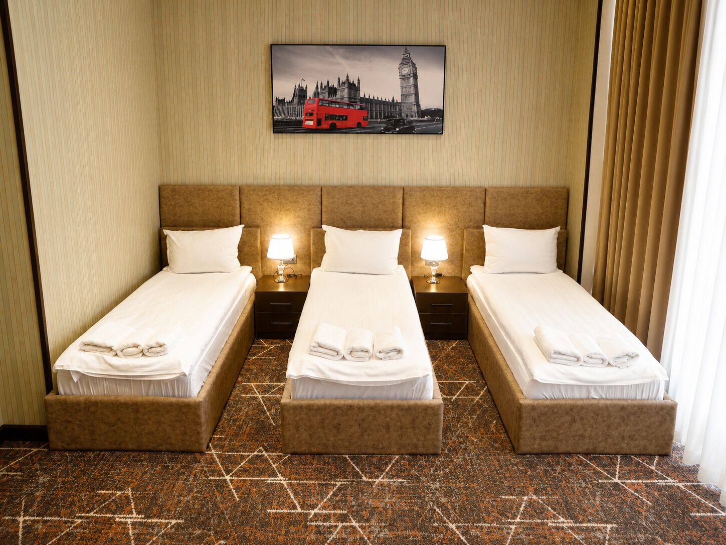 Une chambre d’hôtel avec deux lits simples, une tête de lit, des lampes de chevet, un tableau représentant un autobus à impériale rouge accroché au mur, et une moquette à motifs.