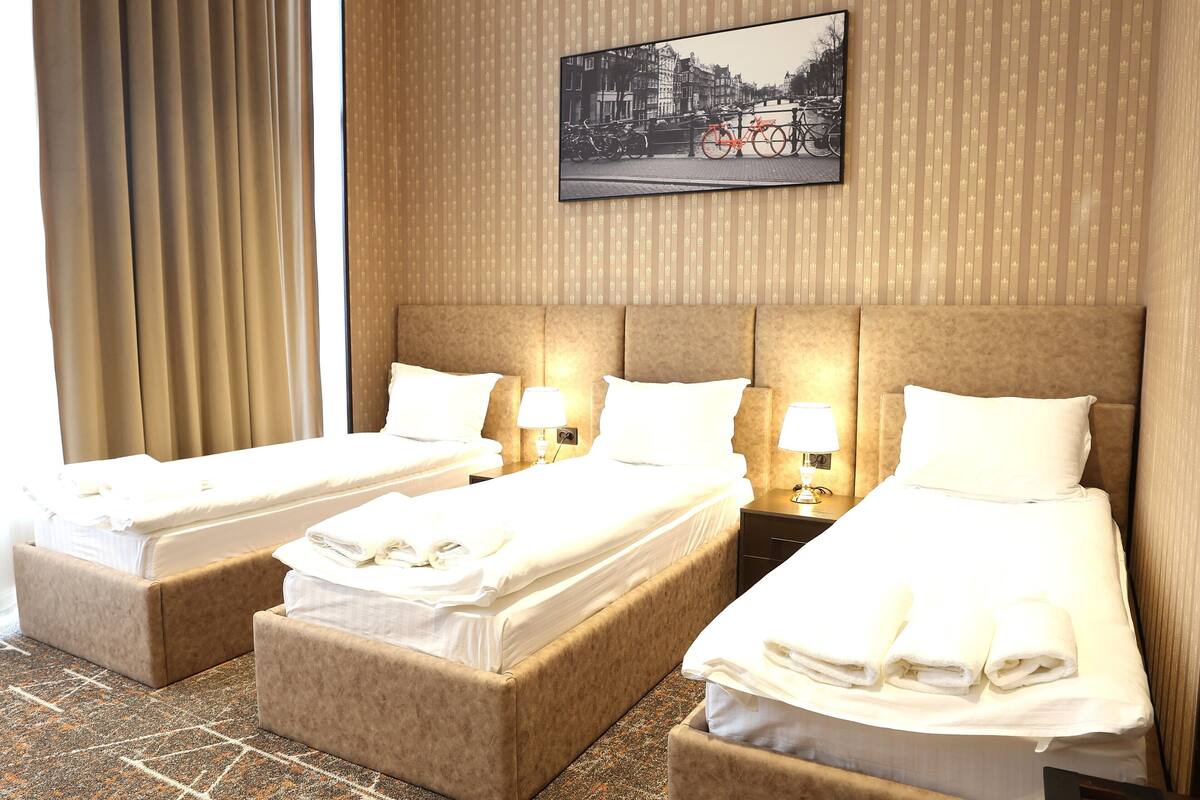 Une chambre d’hôtel avec trois lits, une grande fenêtre avec des rideaux, un tableau accroché au mur et un papier peint à motifs.
